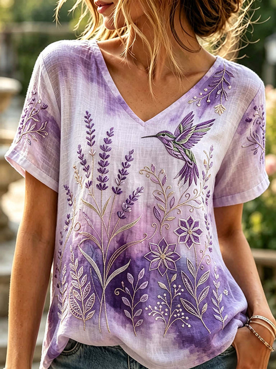 A woman wearing a short-sleeve V-neck top with a purple and lavender watercolor wash, decorated with embroidered lavender stems, flowers, and a hummingbird in green and purple.