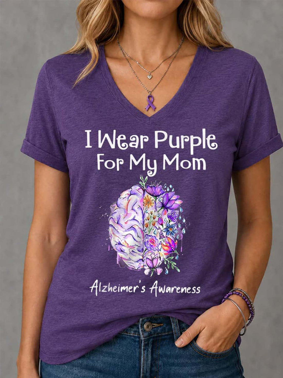 A woman wearing a heather purple V-neck t-shirt with a graphic that reads 'I Wear Purple For My Mom' and 'Alzheimer's Awareness' above a watercolor brain illustration. The background is a neutral gray wall.