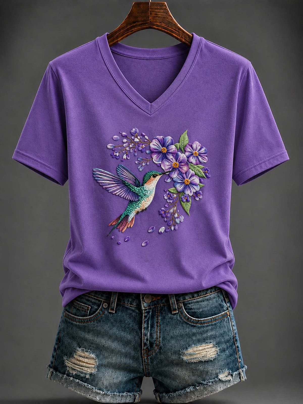 A purple short-sleeve V-neck T-shirt with a colorful hummingbird and purple flowers graphic on the front, styled with distressed denim shorts on a hanger against a dark gray background.