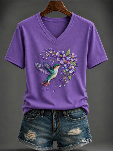 A purple short-sleeve V-neck T-shirt with a colorful hummingbird and purple flowers graphic on the front, styled with distressed denim shorts on a hanger against a dark gray background.