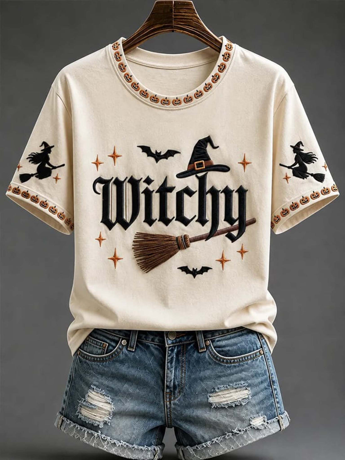 Cream-colored short-sleeve t-shirt with a Halloween 'Witchy' graphic print, styled with distressed denim shorts on a hanger against a dark gray background.