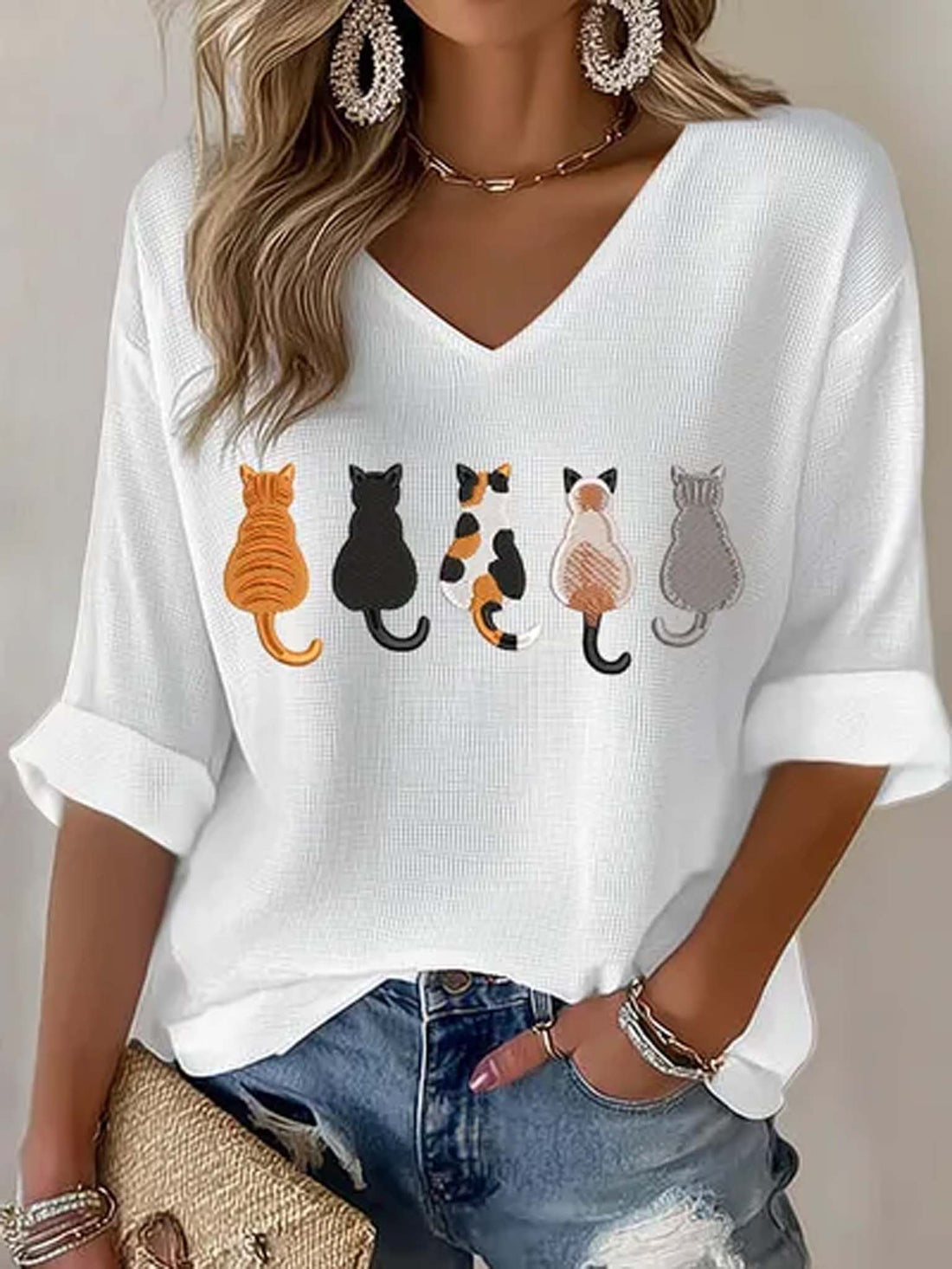 A woman wearing a white V-neck knit top with a row of colorful cat illustrations across the chest, paired with blue jeans and jewelry.