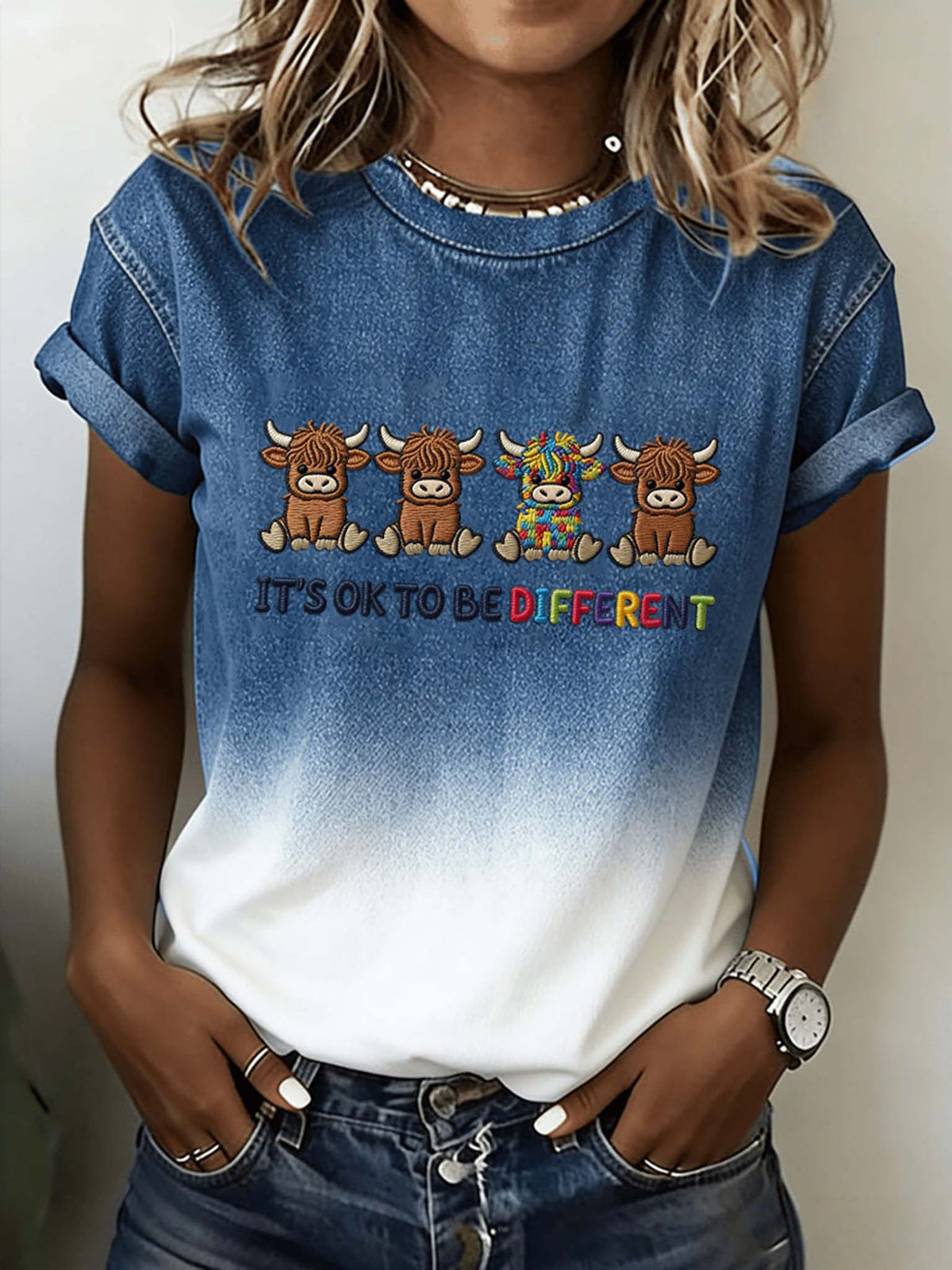 A woman wearing a blue-to-white gradient t-shirt with a graphic of four Scottish Highland cows and the text 'IT'S OK TO BE DIFFERENT' in rainbow colors.
