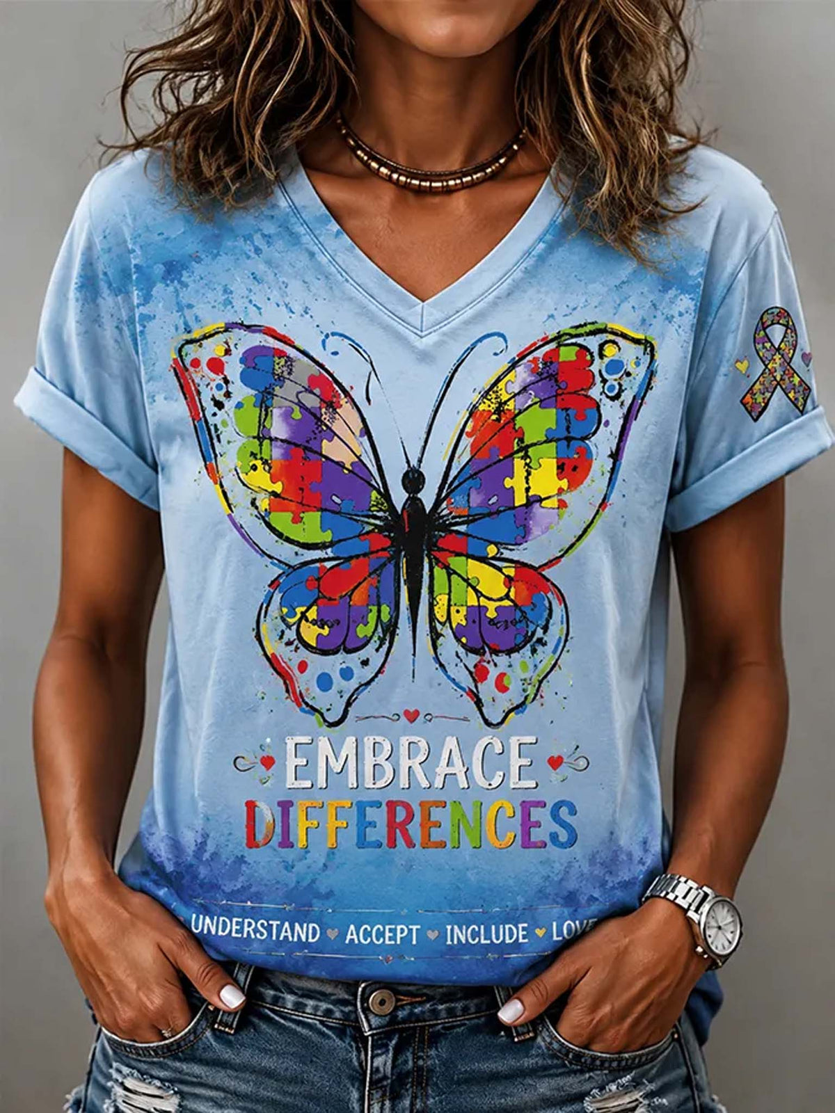 A woman wearing a light blue V-neck graphic T-shirt with a large colorful butterfly print, the words 'EMBRACE DIFFERENCES,' and a small awareness ribbon on the sleeve, paired with blue jeans.