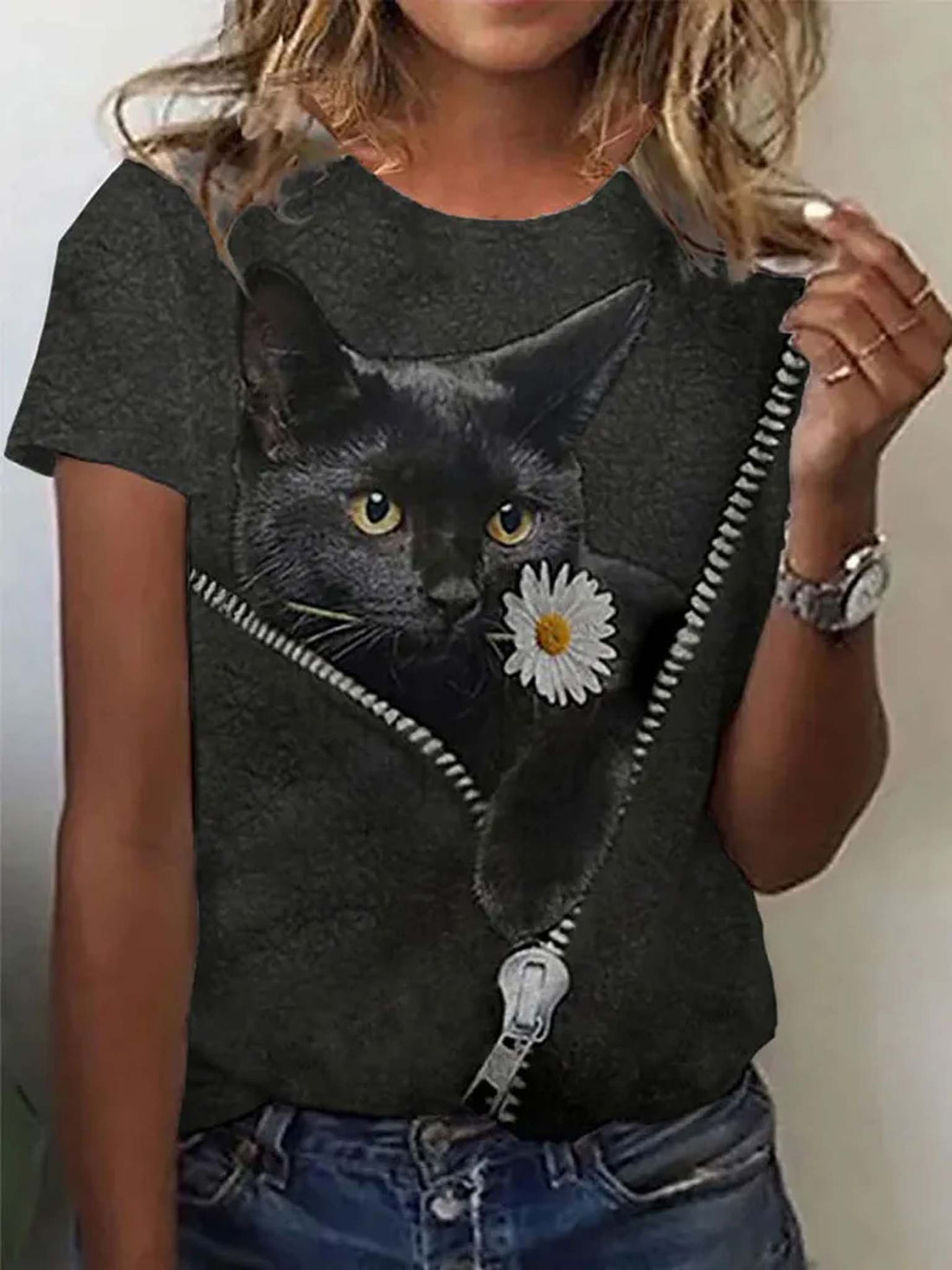 A woman wearing a dark gray t-shirt with a graphic of a black cat peeking out from a zipper, holding a white daisy flower. The background is a plain light wall.