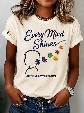 A woman wearing a cream-colored graphic t-shirt with the text 'Every Mind Shines' and 'AUTISM ACCEPTANCE' alongside a silhouette and colorful puzzle pieces, paired with blue jeans.