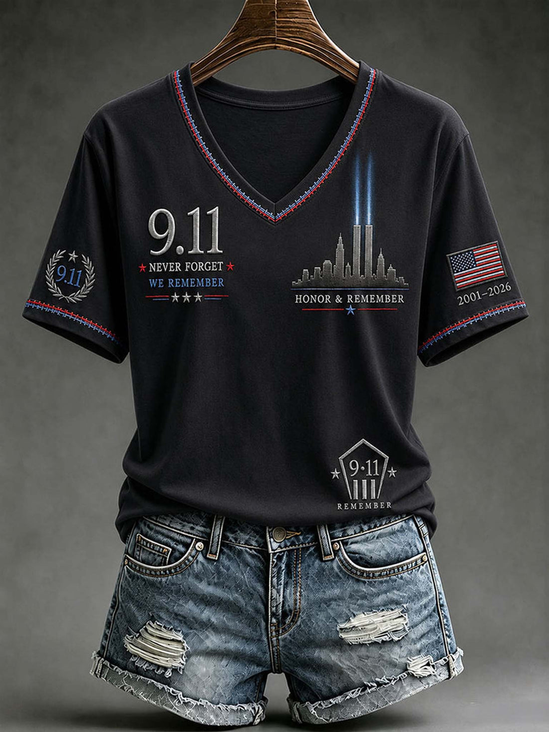 A black V-neck t-shirt with 9/11 themed graphics, shown on a wooden hanger with distressed denim shorts against a dark gray studio background.