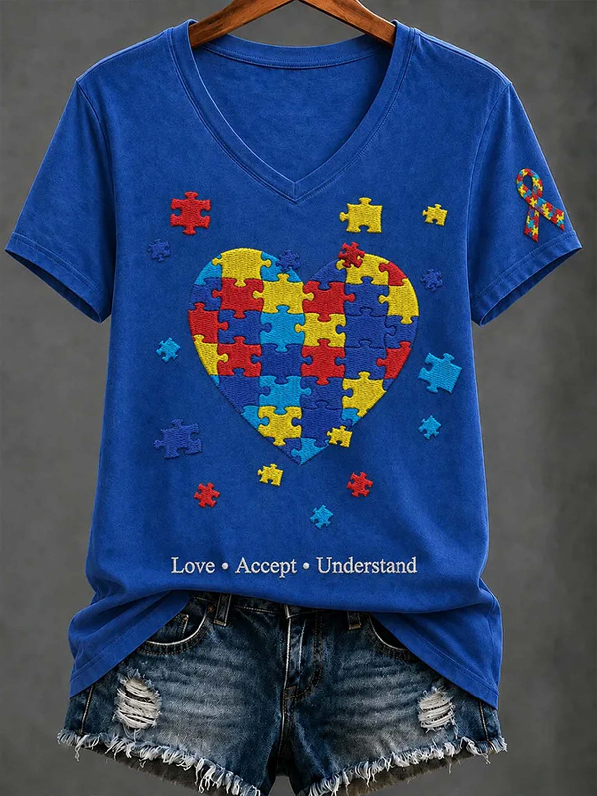 A royal blue V-neck t-shirt on a wooden hanger, styled with distressed denim shorts. The shirt features a large heart-shaped autism awareness puzzle graphic in the center, surrounded by small colorful puzzle pieces, with the text 'Love • Accept • Understand' printed at the bottom.