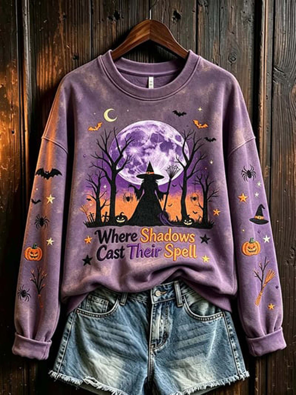A lavender-purple crewneck sweatshirt with a Halloween graphic print, styled on a wooden hanger with denim shorts against a dark wood plank background. The graphic features a witch silhouette, a full moon, bats, pumpkins, and the text 'Where Shadows Cast Their Spell'.