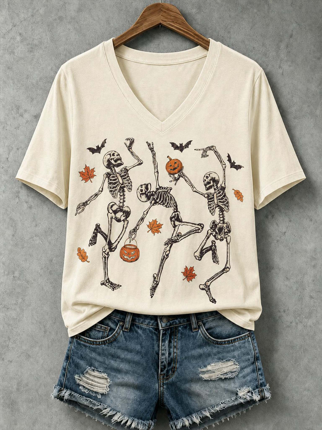 An off-white V-neck t-shirt with a graphic of three dancing skeletons, pumpkins, bats, and autumn leaves, styled on a wooden hanger with distressed denim shorts against a textured gray background.